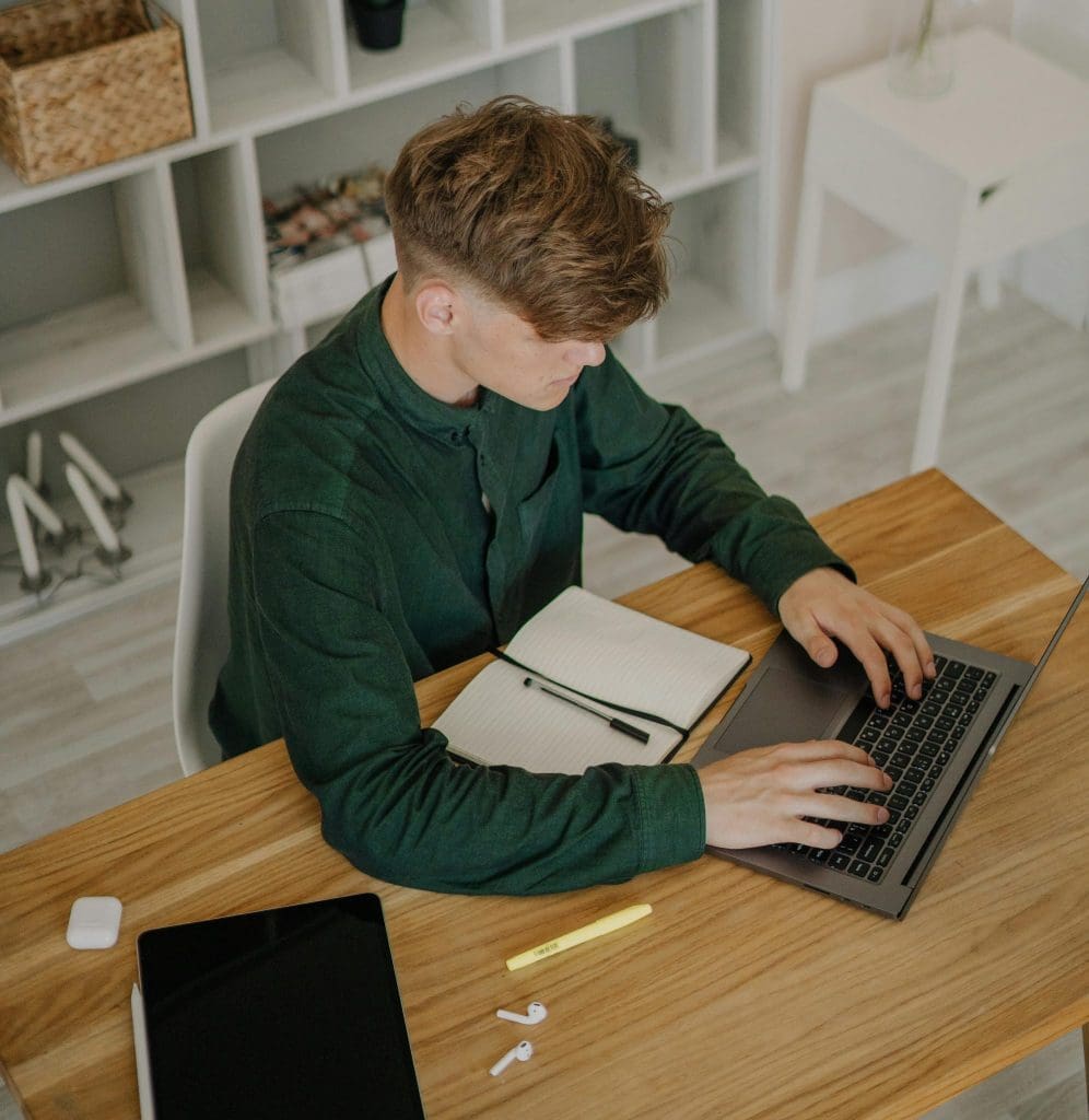 A young man with light hair wearing a forest green long-sleeved shirt using a laptop at a desk