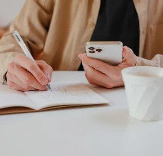 A person slightly off-screen holding a phone and writing in a notepad